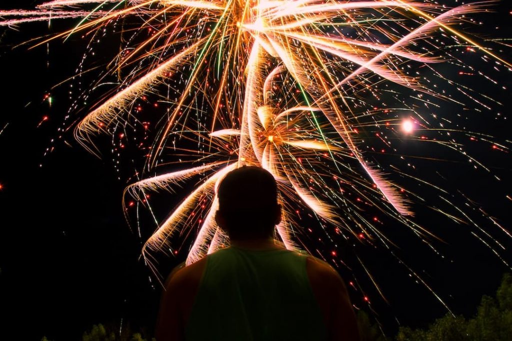 A man in a tank top stands watching a bright fireworks display